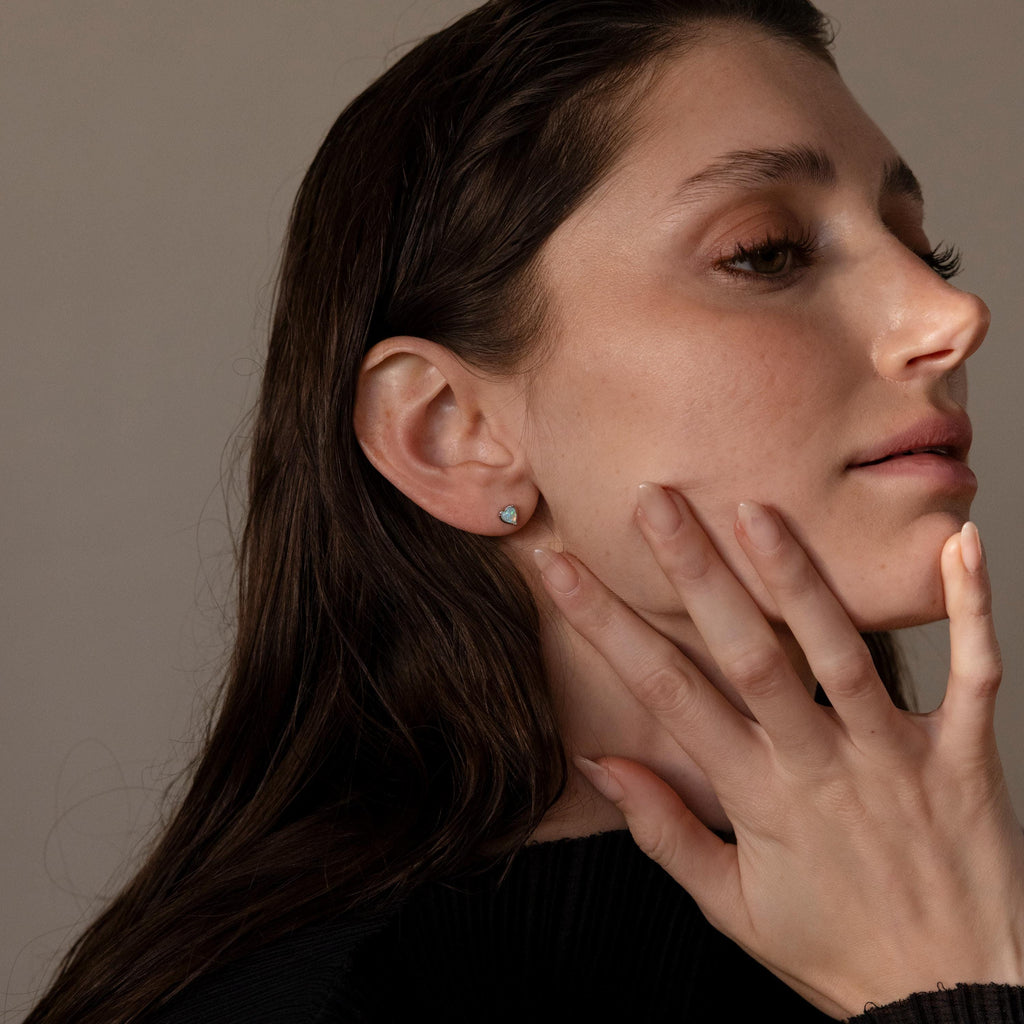 A woman with long brown hair touches her face, wearing a black top and Opal Heart Flat Back Studs that add a subtle sparkle to her look.