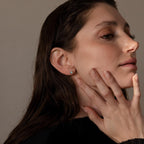 A woman with long brown hair touches her face, wearing a black top and Opal Heart Flat Back Studs that add a subtle sparkle to her look.