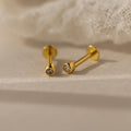 The Small Diamond Bezel Flatback Studs, featuring clear gemstones, rest on a soft beige surface near white fabric, highlighting the timeless elegance of classic diamond earrings.