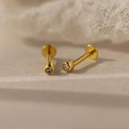 The Small Diamond Bezel Flatback Studs, featuring clear gemstones, rest on a soft beige surface near white fabric, highlighting the timeless elegance of classic diamond earrings.