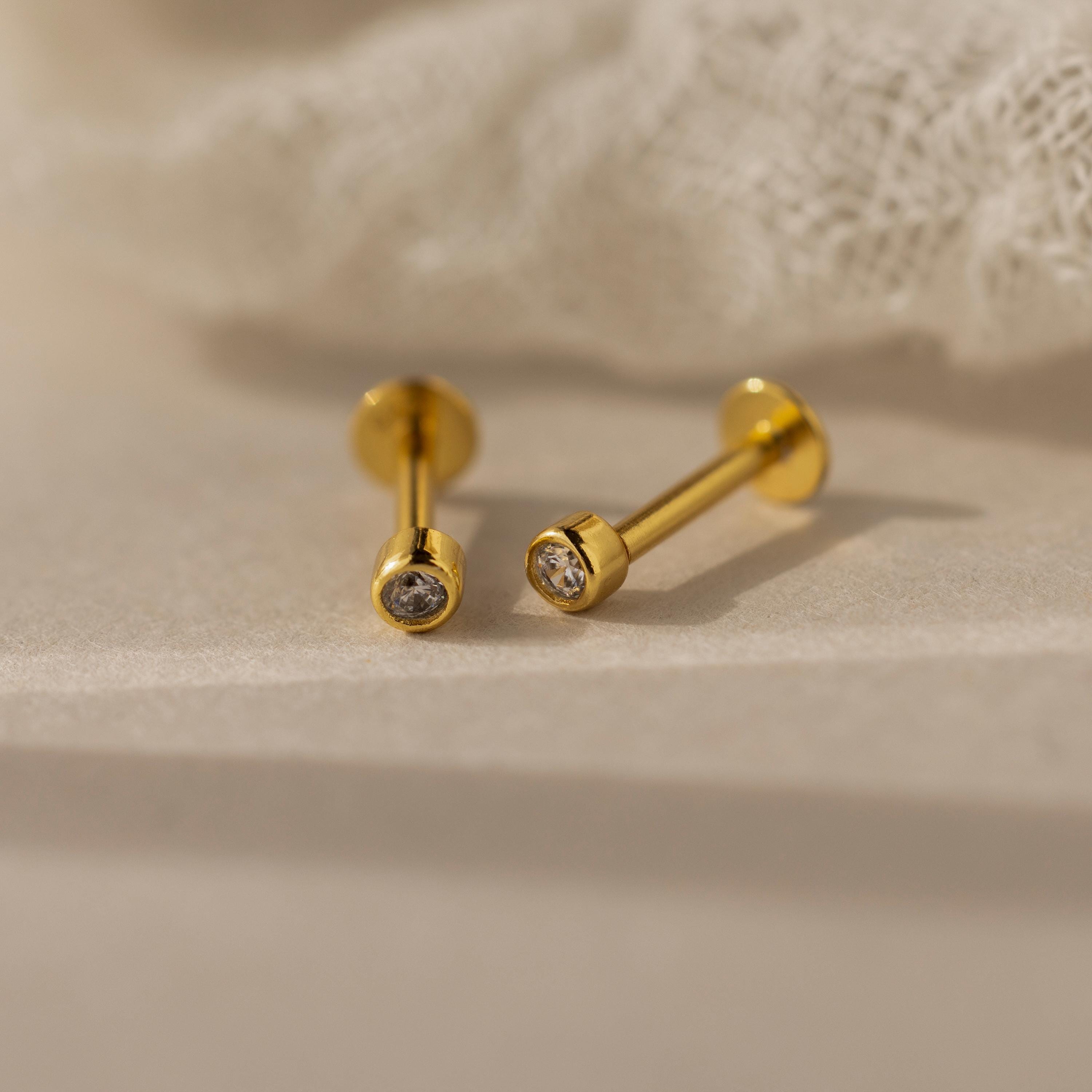 The Small Diamond Bezel Flatback Studs, featuring clear gemstones, rest on a soft beige surface near white fabric, highlighting the timeless elegance of classic diamond earrings.