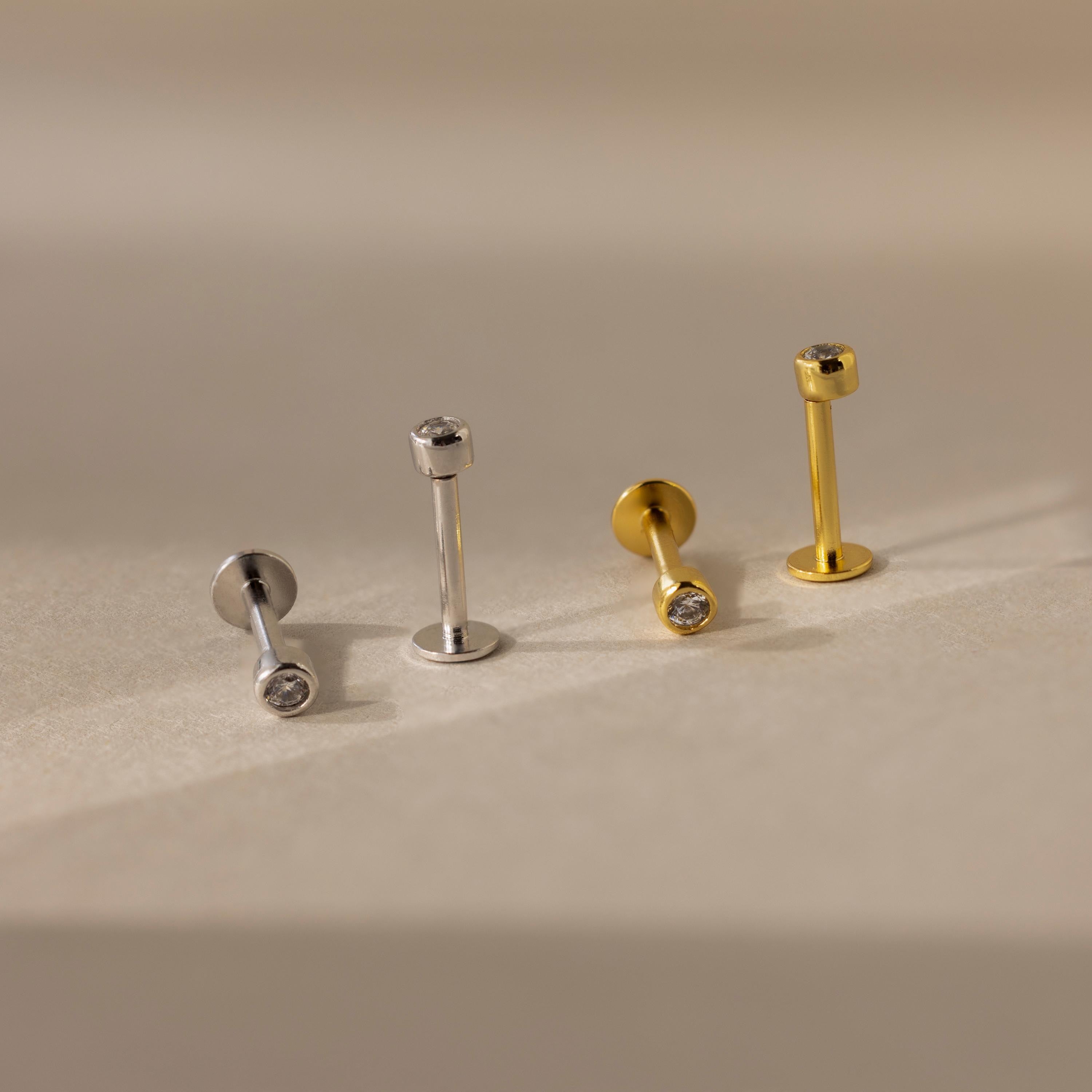 Two pairs of Small Diamond Bezel Flatback Studs—one in silver and one in gold, each with clear stones—are displayed on a beige surface.