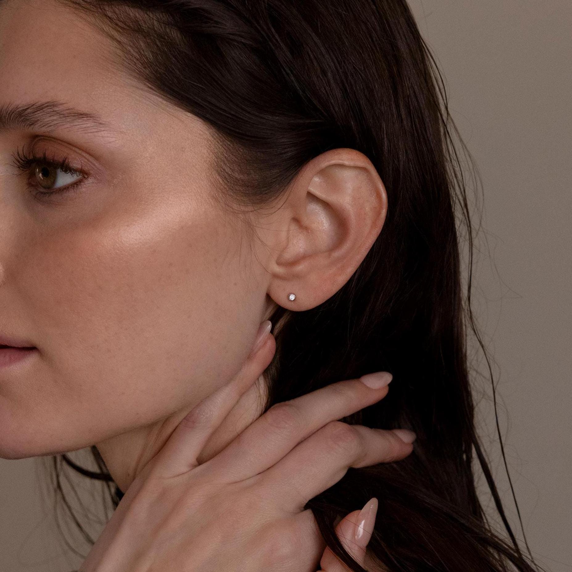 A woman with dewy skin wears Small Diamond Bezel Flatback Studs and touches her neck with manicured fingers.