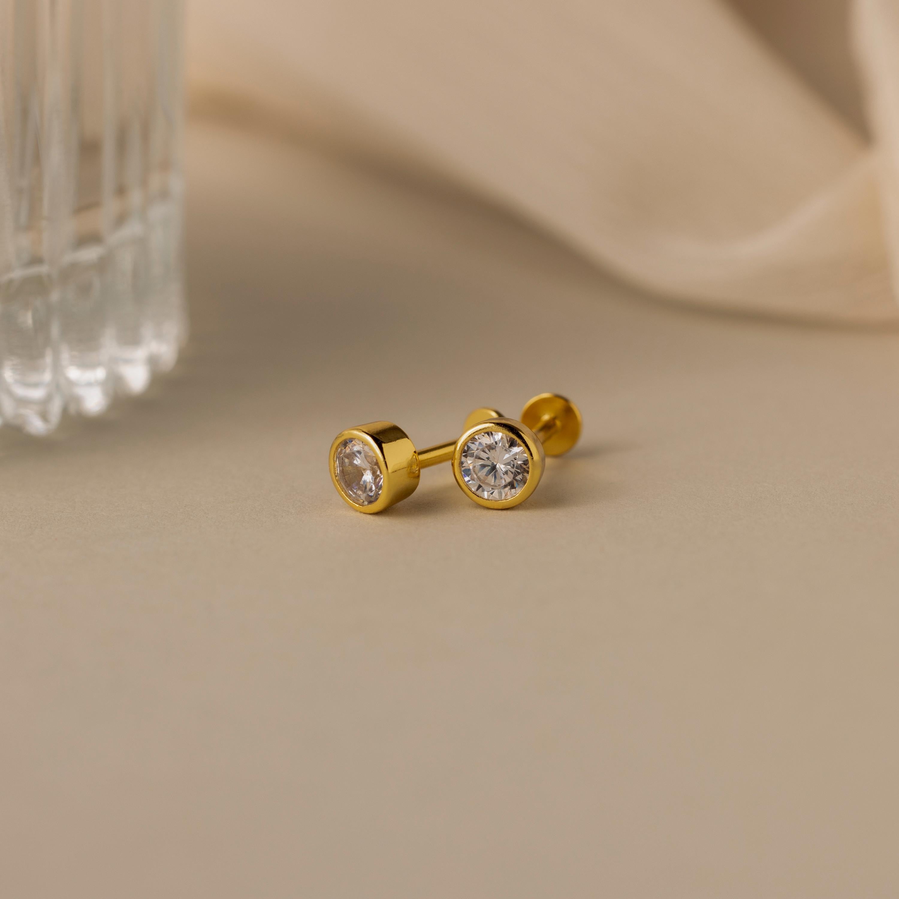 Two Large Diamond Bezel Flatback Studs are displayed on a beige surface near a glass object.