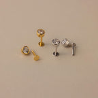 Two pairs of Large Diamond Bezel Flatback Studs—one gold, one silver—each with a round clear gemstone, evoke classic diamond studs and are displayed on a beige background.