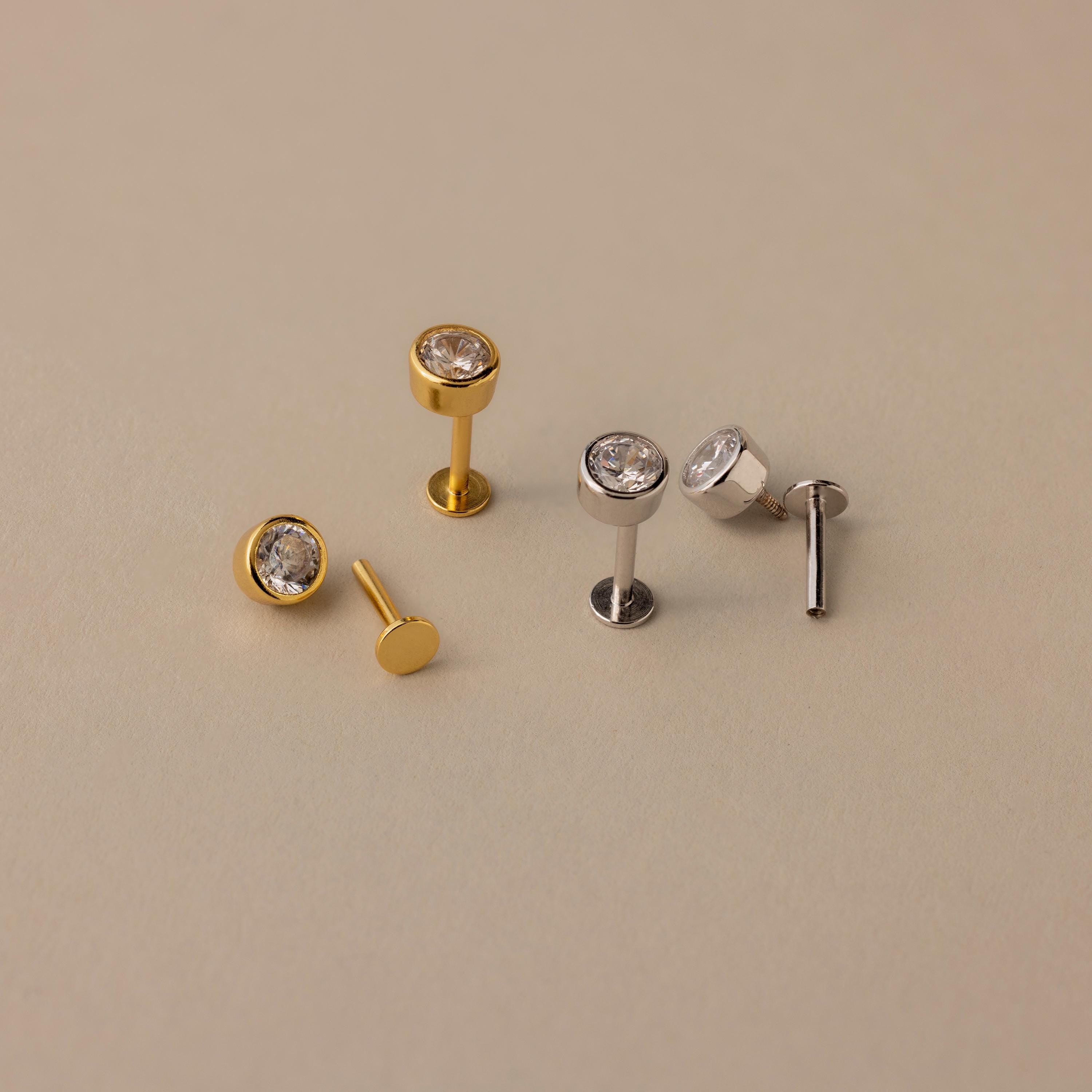 Two pairs of Large Diamond Bezel Flatback Studs—one gold, one silver—each with a round clear gemstone, evoke classic diamond studs and are displayed on a beige background.