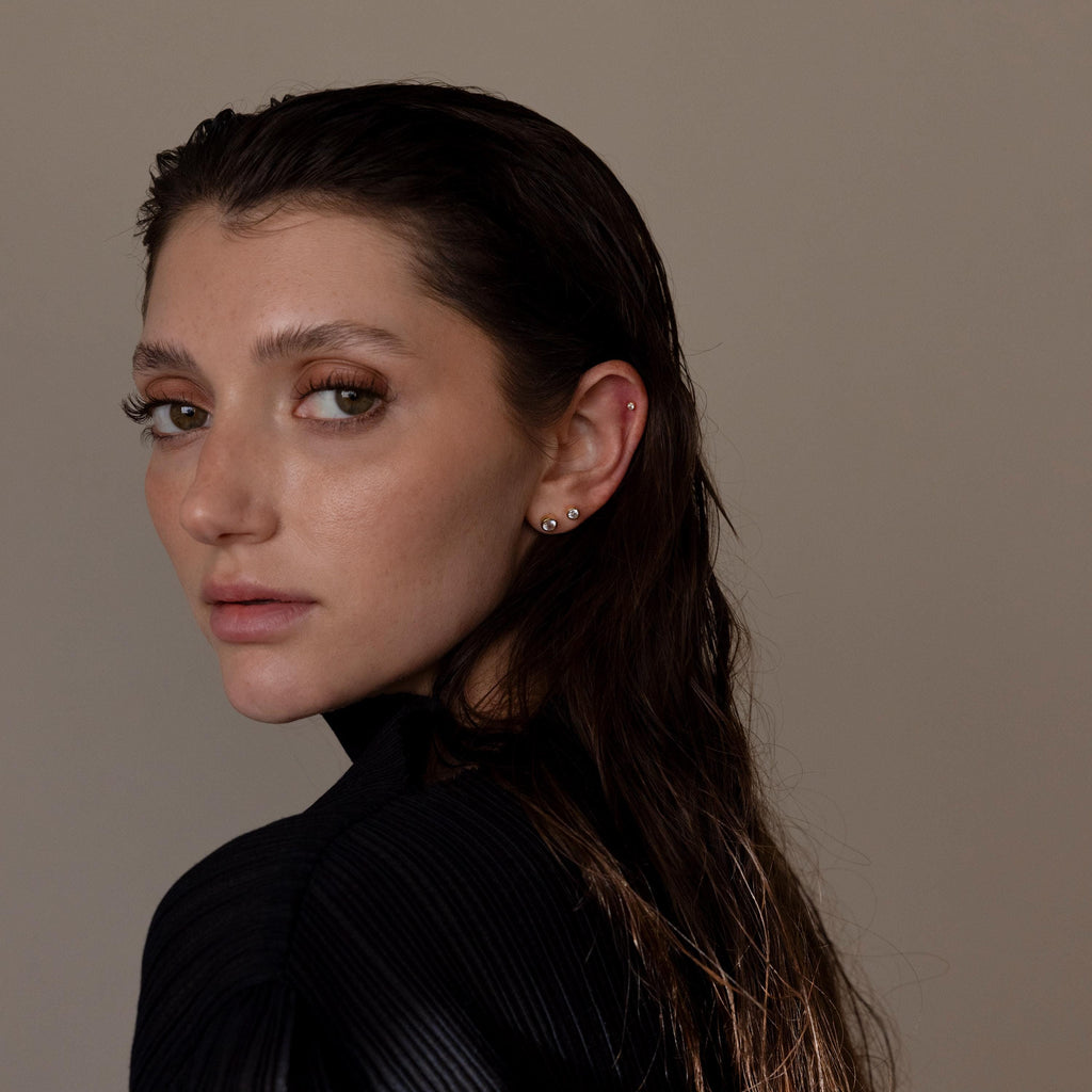 A woman with wet dark hair and several ear piercings, including Large Diamond Bezel Flatback Studs, looks over her shoulder against a plain backdrop.