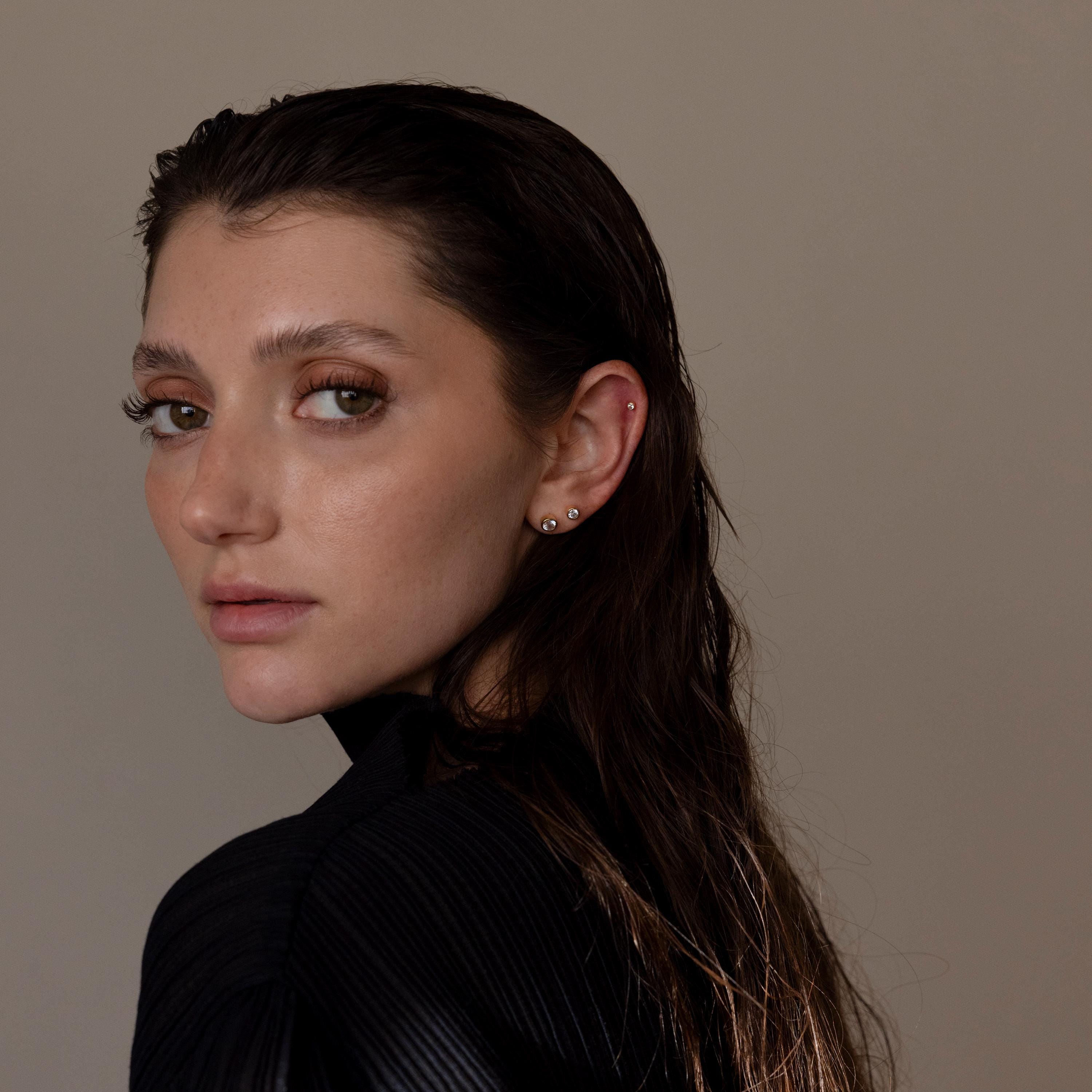 A woman with wet dark hair and several ear piercings, including Large Diamond Bezel Flatback Studs, looks over her shoulder against a plain backdrop.