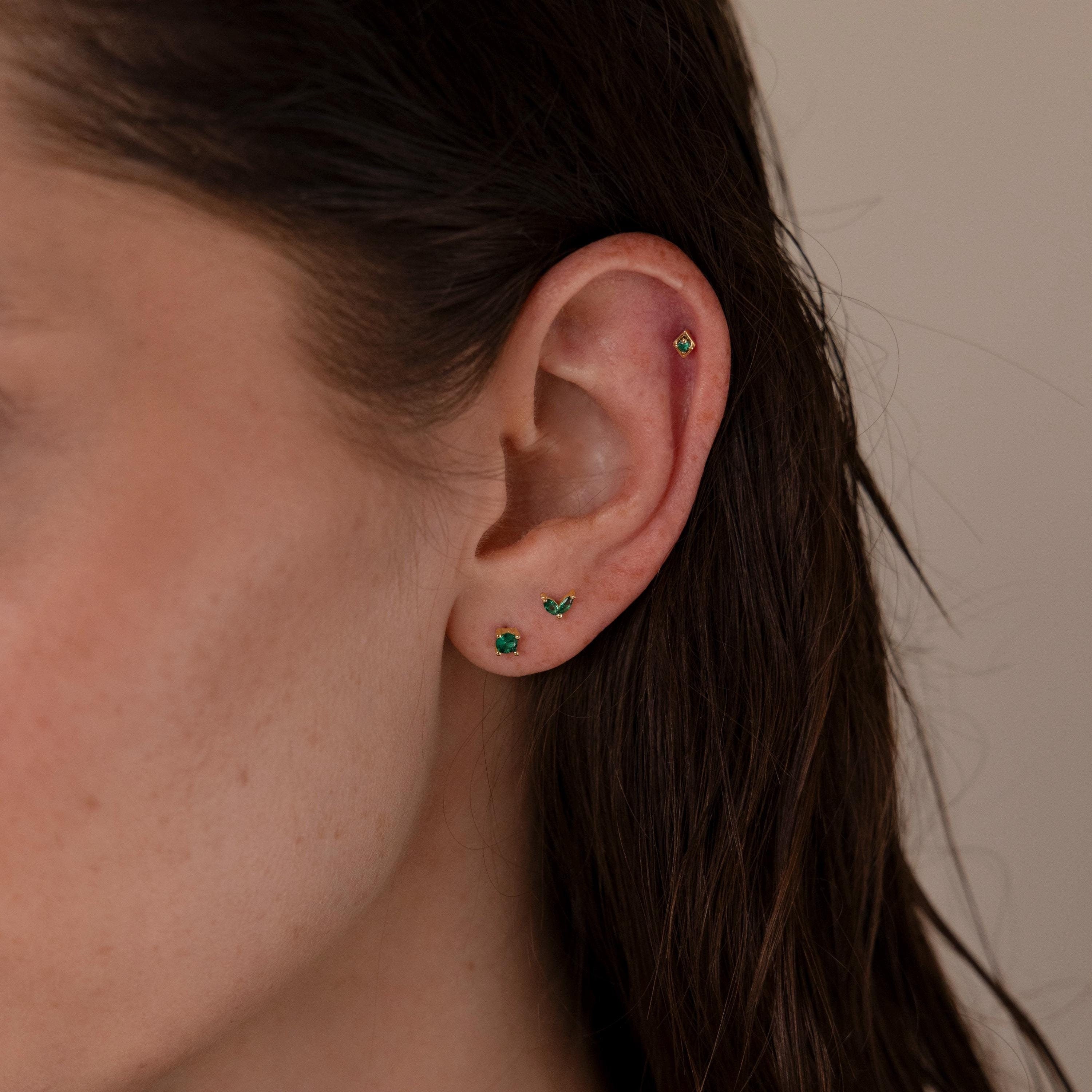 Close-up of a woman's ear wearing the Emerald Flat Back Earring Set, featuring three emerald earrings with birthstone studs on the lobe and a screw-back earring on the upper cartilage.