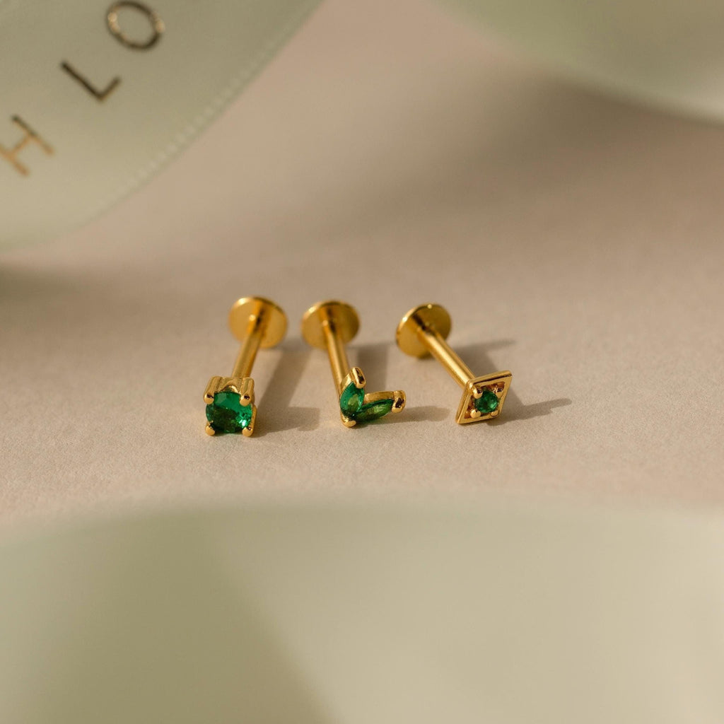 The Emerald Flat Back Earring Set features three gold stud earrings with green gemstones—heart, round, and square—elegantly arranged on a beige surface. Perfect as birthstone studs for any occasion.