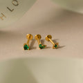 The Emerald Flat Back Earring Set features three gold stud earrings with green gemstones—heart, round, and square—elegantly arranged on a beige surface. Perfect as birthstone studs for any occasion.