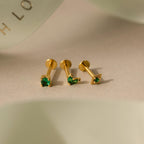 The Emerald Flat Back Earring Set features three gold stud earrings with green gemstones—heart, round, and square—elegantly arranged on a beige surface. Perfect as birthstone studs for any occasion.