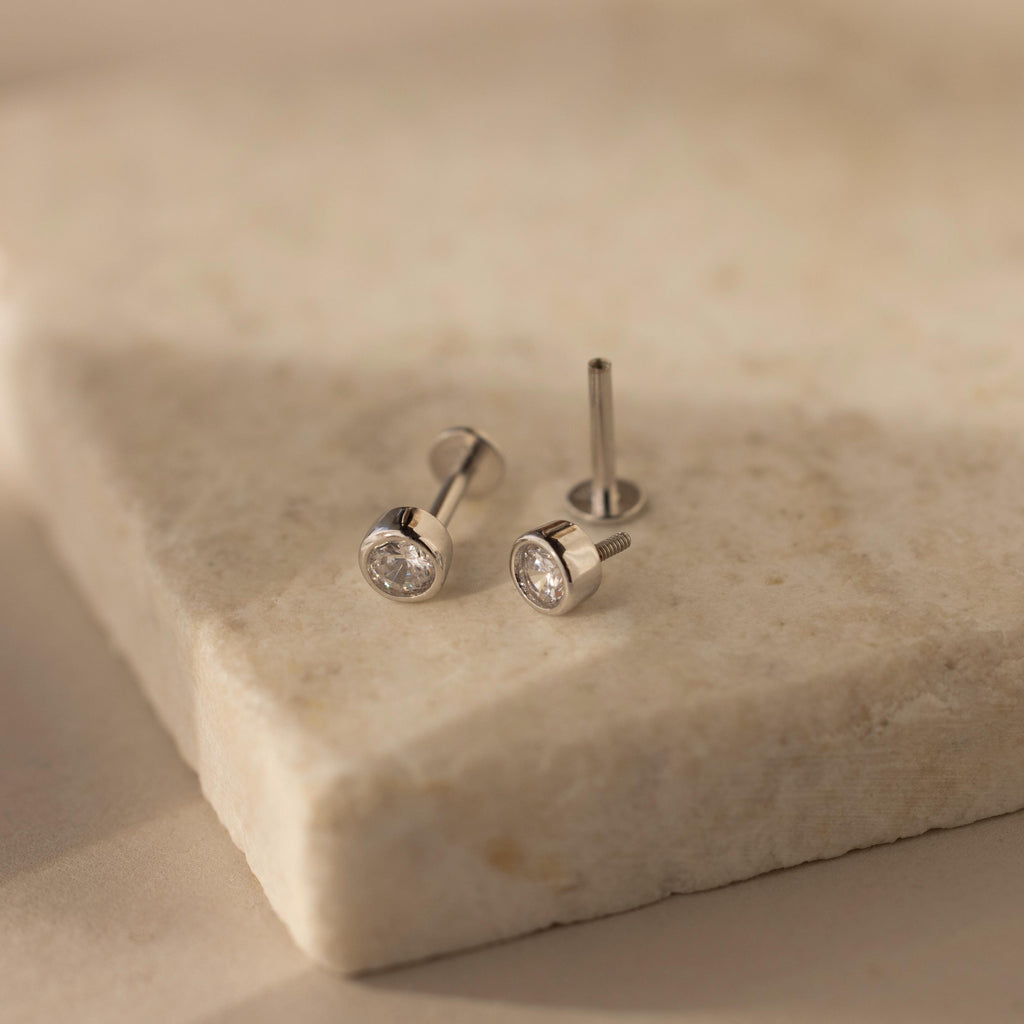 Showcased on a light stone surface, the Medium Diamond Bezel Flatback Studs feature silver settings with round clear gemstones—an elegant addition to any jewelry collection.
