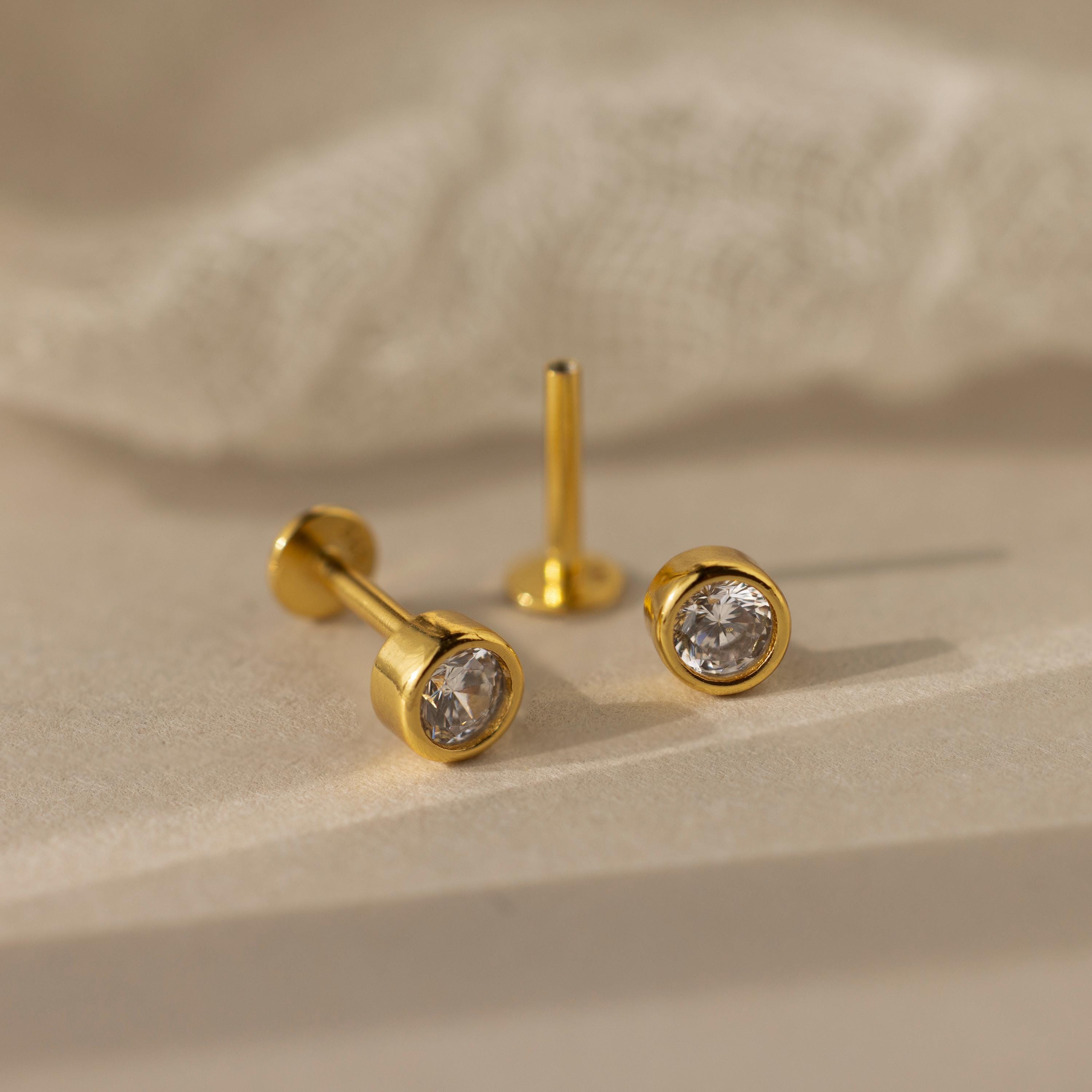 The Medium Diamond Bezel Flatback Studs, a pair of gold earrings with clear gemstones, are perfect for any jewelry collection and elegantly displayed on a beige surface with a soft fabric backdrop.