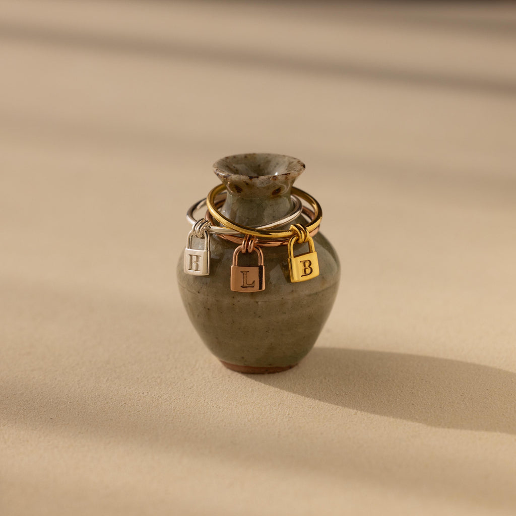 The Initial Padlock Drop Ring is a small ceramic vase featuring three metal initial charms—H, L, and B—dangling around its neck, evoking the look of personalized jewelry or an initial padlock ring.