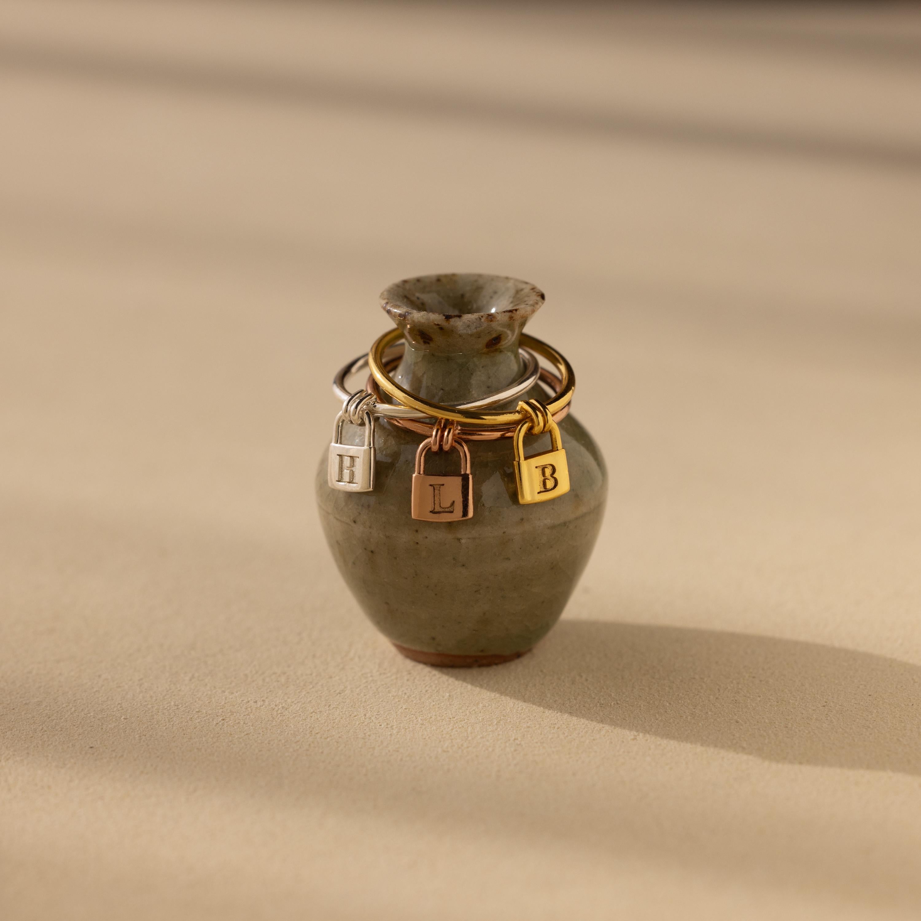 The Initial Padlock Drop Ring is a small ceramic vase featuring three metal initial charms—H, L, and B—dangling around its neck, evoking the look of personalized jewelry or an initial padlock ring.
