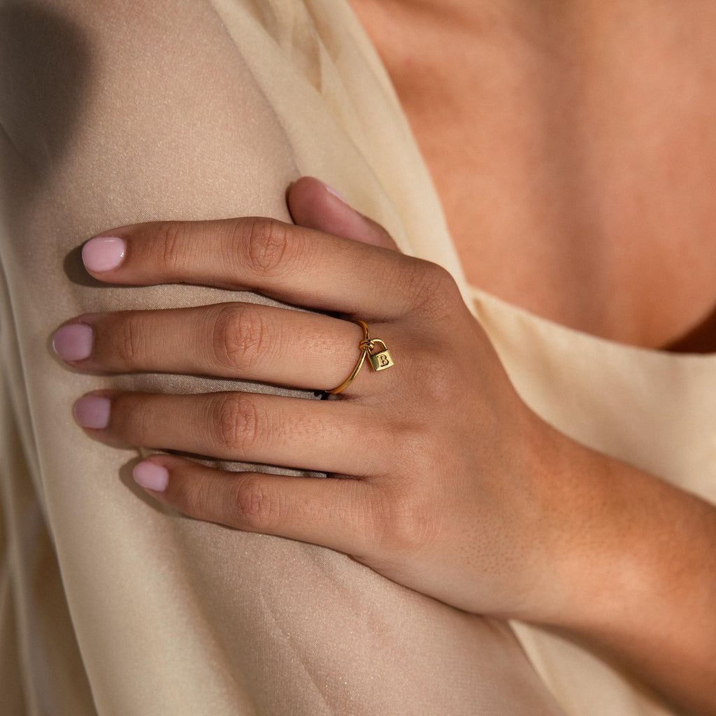 A hand with pale pink nails wears the Initial Padlock Drop Ring—a gold ring with a lock charm—resting on light beige fabric, perfect for fans of personalized jewelry.
