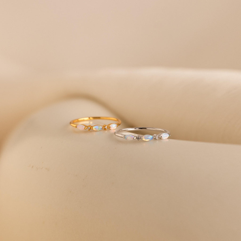 Side-by-side display of the Alessia Trio Opal Ring in gold and silver variations, each showcasing three marquise-cut opals with delicate diamond accents on slim bands, placed on a soft cream surface.