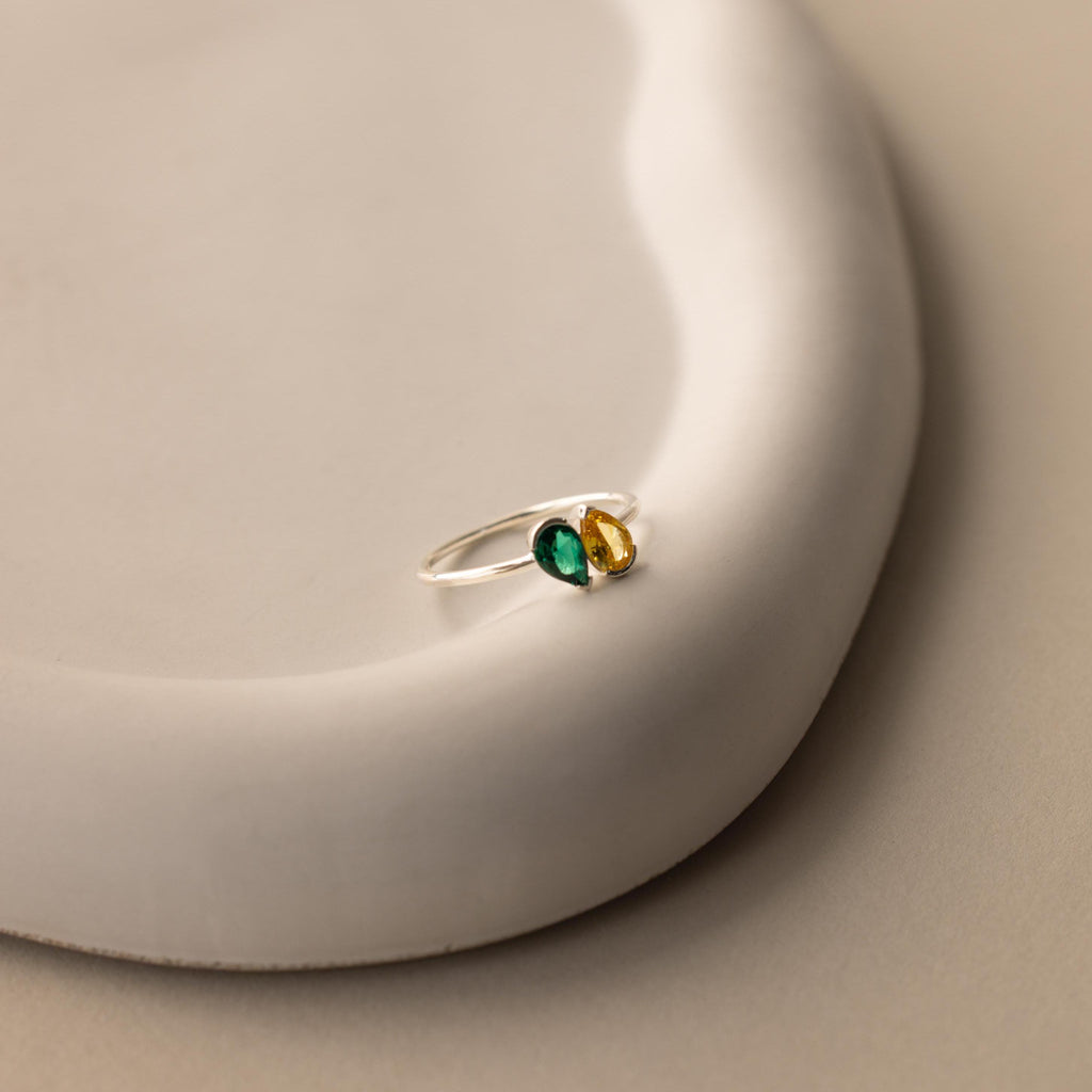 The Toi et Moi Pear Birthstone Ring, featuring green and yellow stones in a silver setting, rests on a smooth, curved white surface—an elegant choice for a romantic gift.