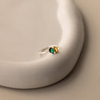 The Toi et Moi Pear Birthstone Ring, featuring green and yellow stones in a silver setting, rests on a smooth, curved white surface—an elegant choice for a romantic gift.