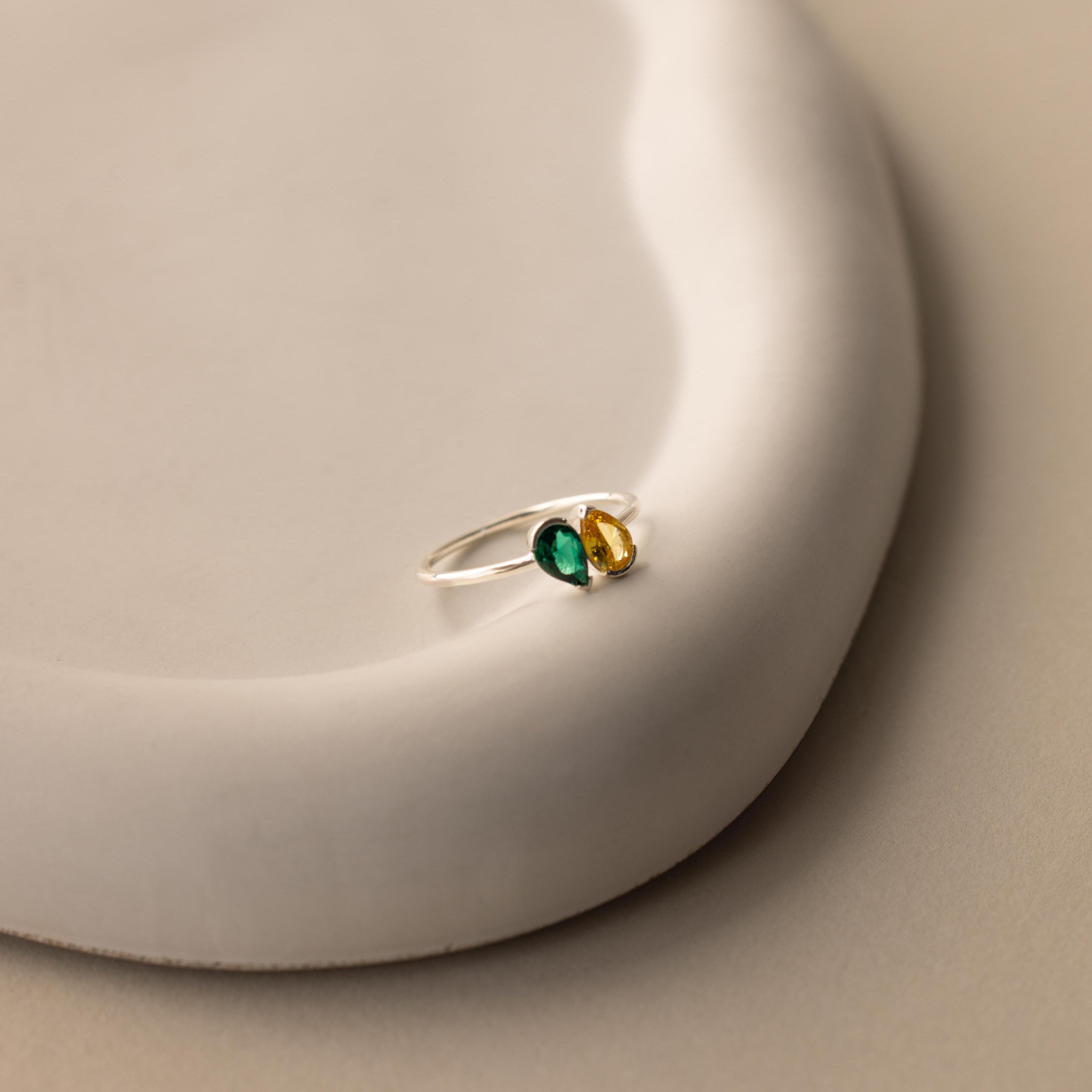 The Toi et Moi Pear Birthstone Ring, featuring green and yellow stones in a silver setting, rests on a smooth, curved white surface—an elegant choice for a romantic gift.