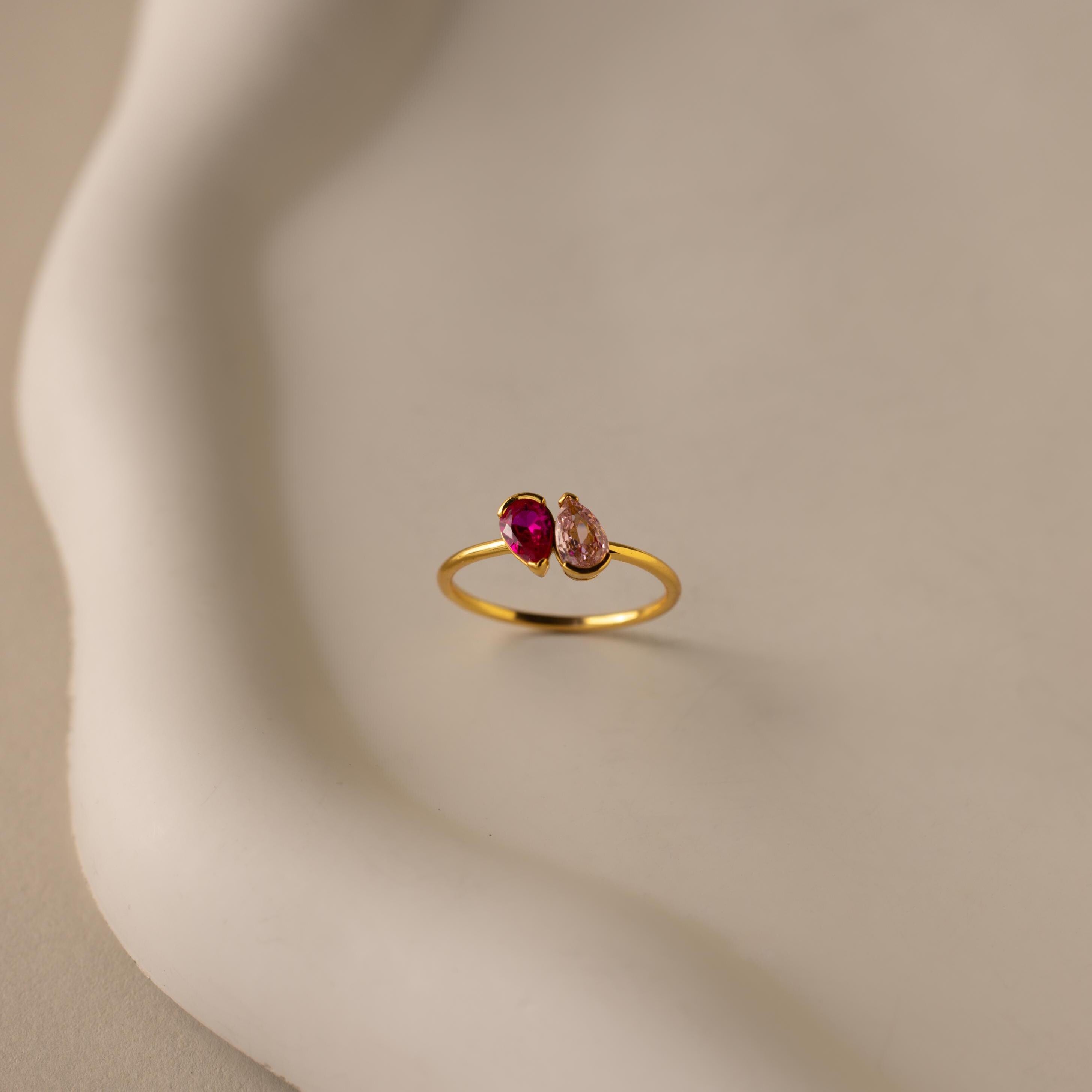 Toi et Moi Pear Birthstone Ring featuring a red and a clear gemstone, displayed on a white, curved ceramic surface—an elegant, romantic gift choice.