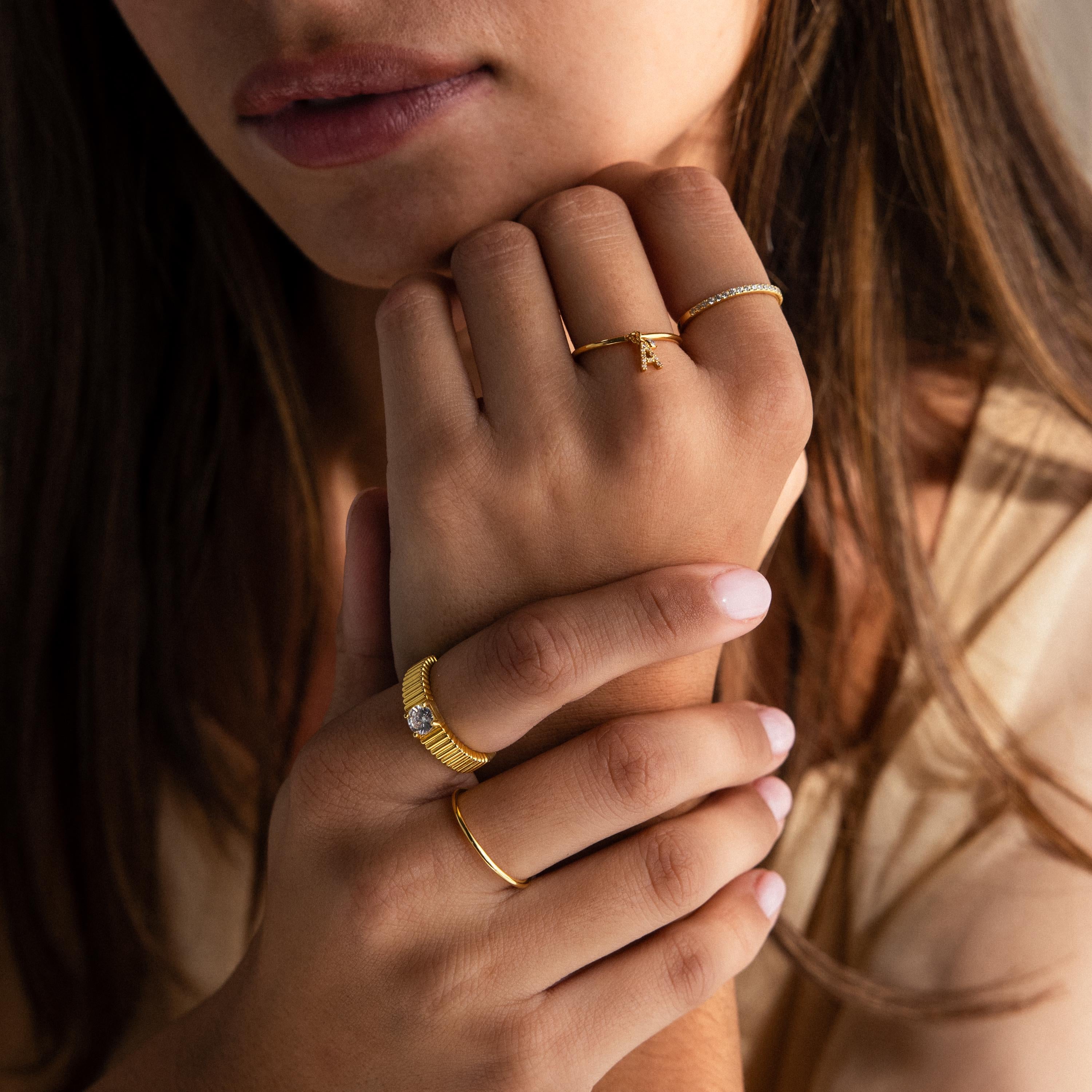 Woman with long brown hair wearing multiple gold rings, including a Pave Initial Birthstone Drop Ring, touching her hands to her face.