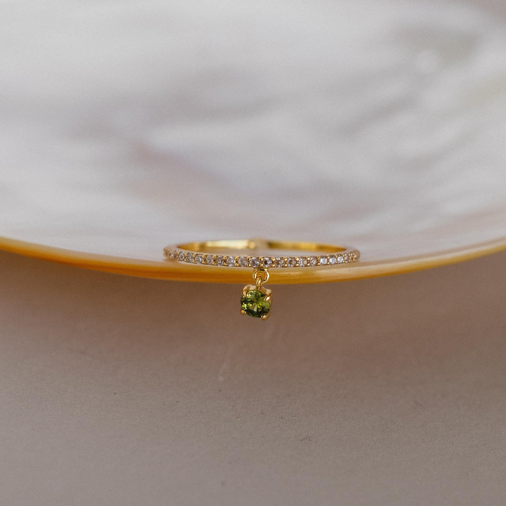 The Pave Birthstone Drop Ring features a gold band with sparkling clear stones in a diamond pave setting and a dangling green gemstone, beautifully displayed on a pearly dish.