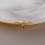 The Pave Birthstone Drop Ring features a gold band with sparkling clear stones in a diamond pave setting and a dangling green gemstone, beautifully displayed on a pearly dish.