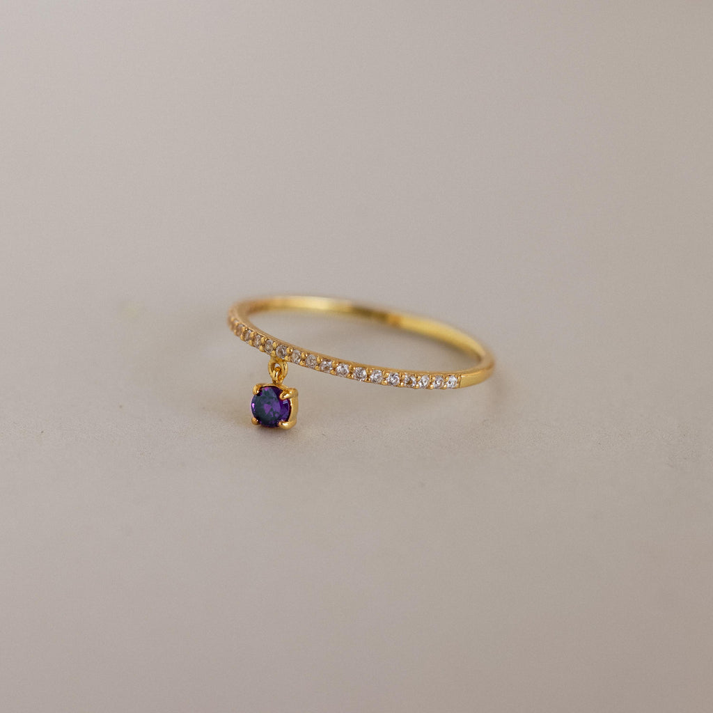 The Pave Birthstone Drop Ring features a delicate gold band with pavé diamonds and a dangling purple birthstone charm, displayed on a neutral background.
