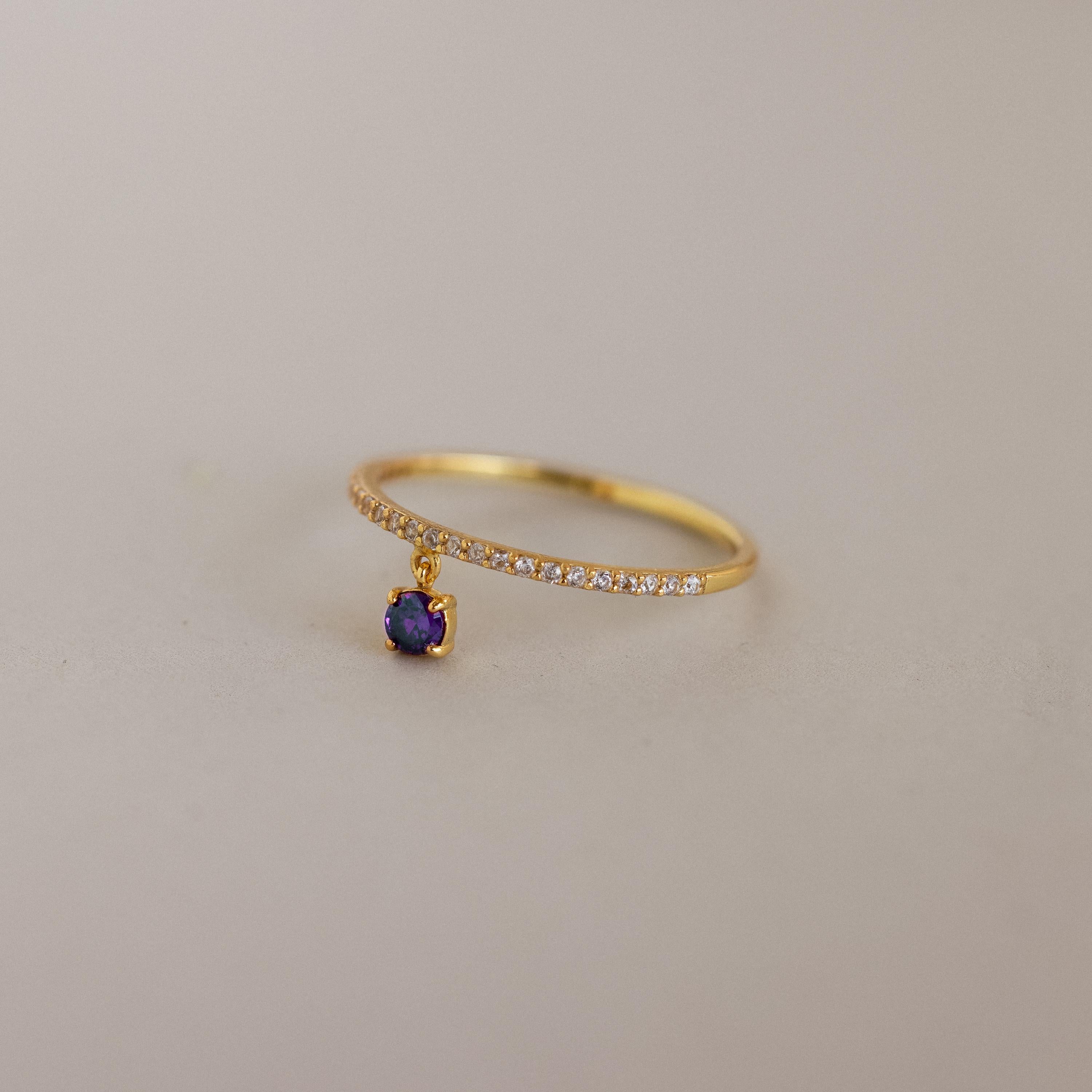 The Pave Birthstone Drop Ring features a delicate gold band with pavé diamonds and a dangling purple birthstone charm, displayed on a neutral background.