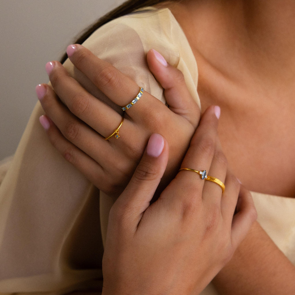 Close-up of hands with light pink nails wearing gold Dainty Birthstone Drop Rings against a beige fabric background—a perfect showcase for personalized jewelry.