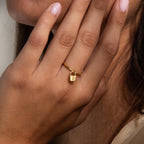 A hand with light pink nails wears an Initial Padlock Drop Ring—a gold ring with a small padlock charm engraved with the letter B, ideal for fans of personalized jewelry.