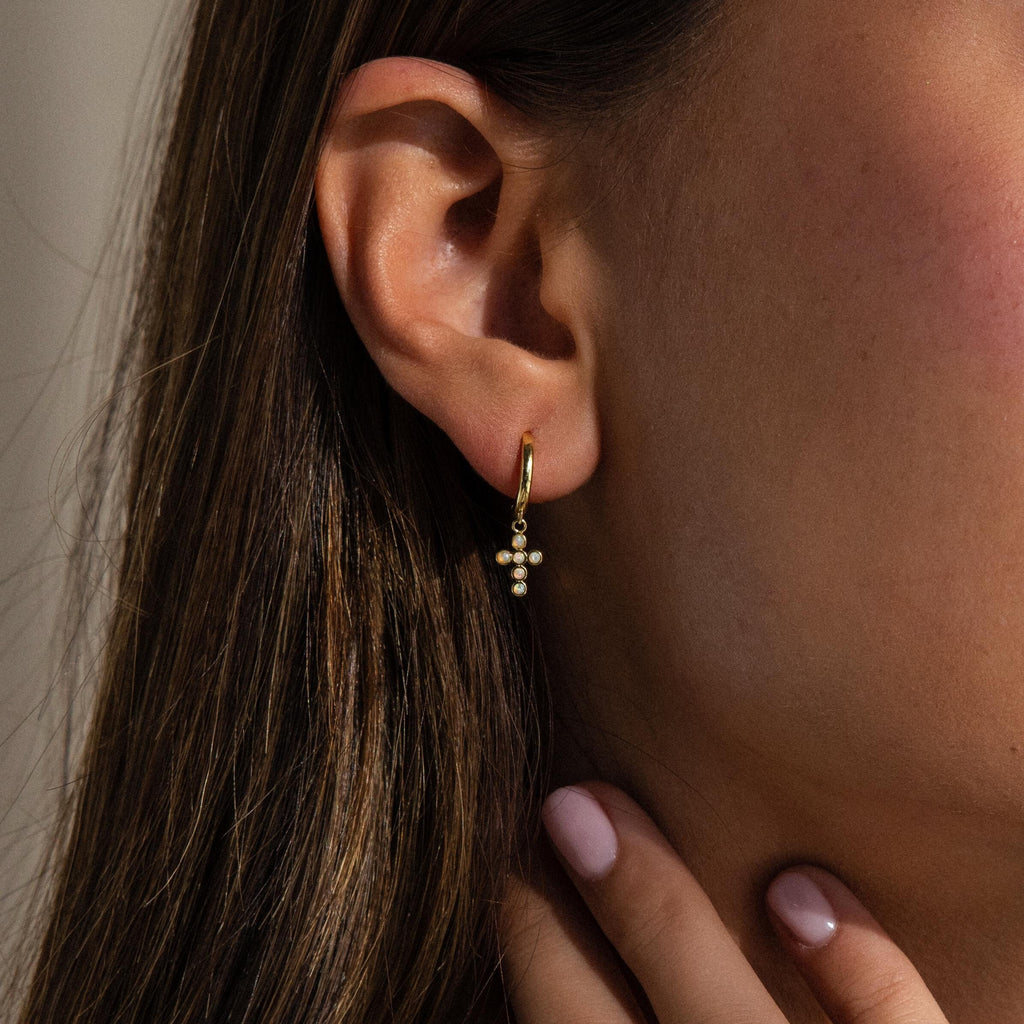 A woman wears Opal Cross Huggies, gently touching her neck with her hand.