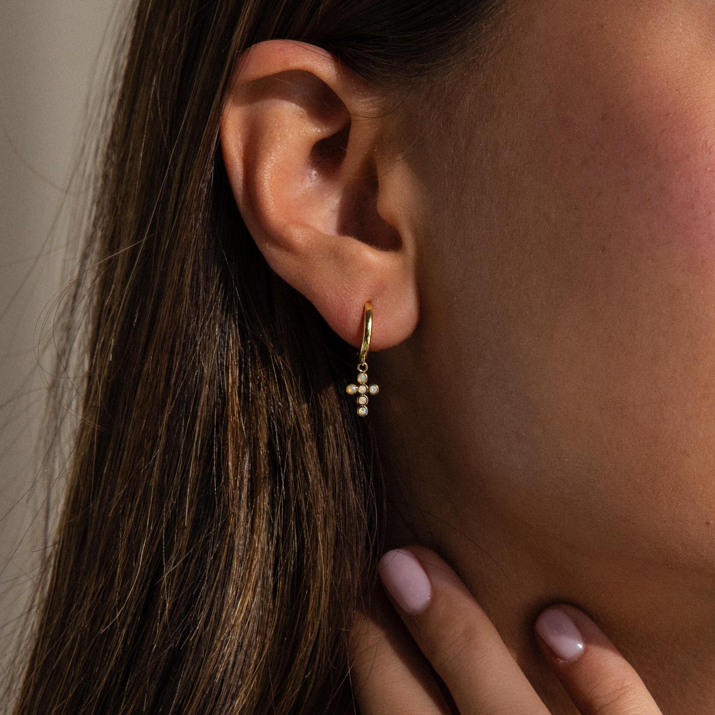 A woman wears Opal Cross Huggies, gently touching her neck with her hand.