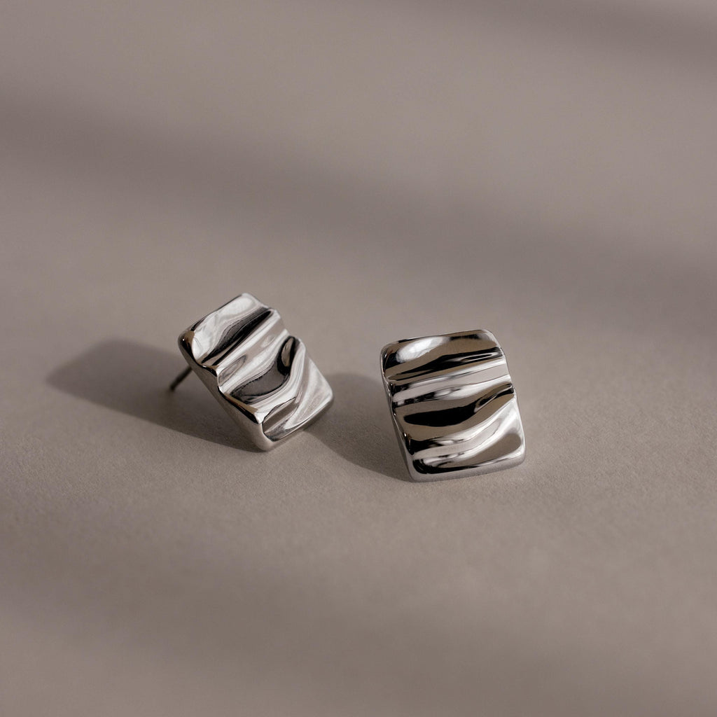 Bold Square Wave Studs in silver feature a wavy, textured surface for a unique touch. Displayed on a neutral background, these minimalist earrings add modern elegance to any luxury jewelry collection.