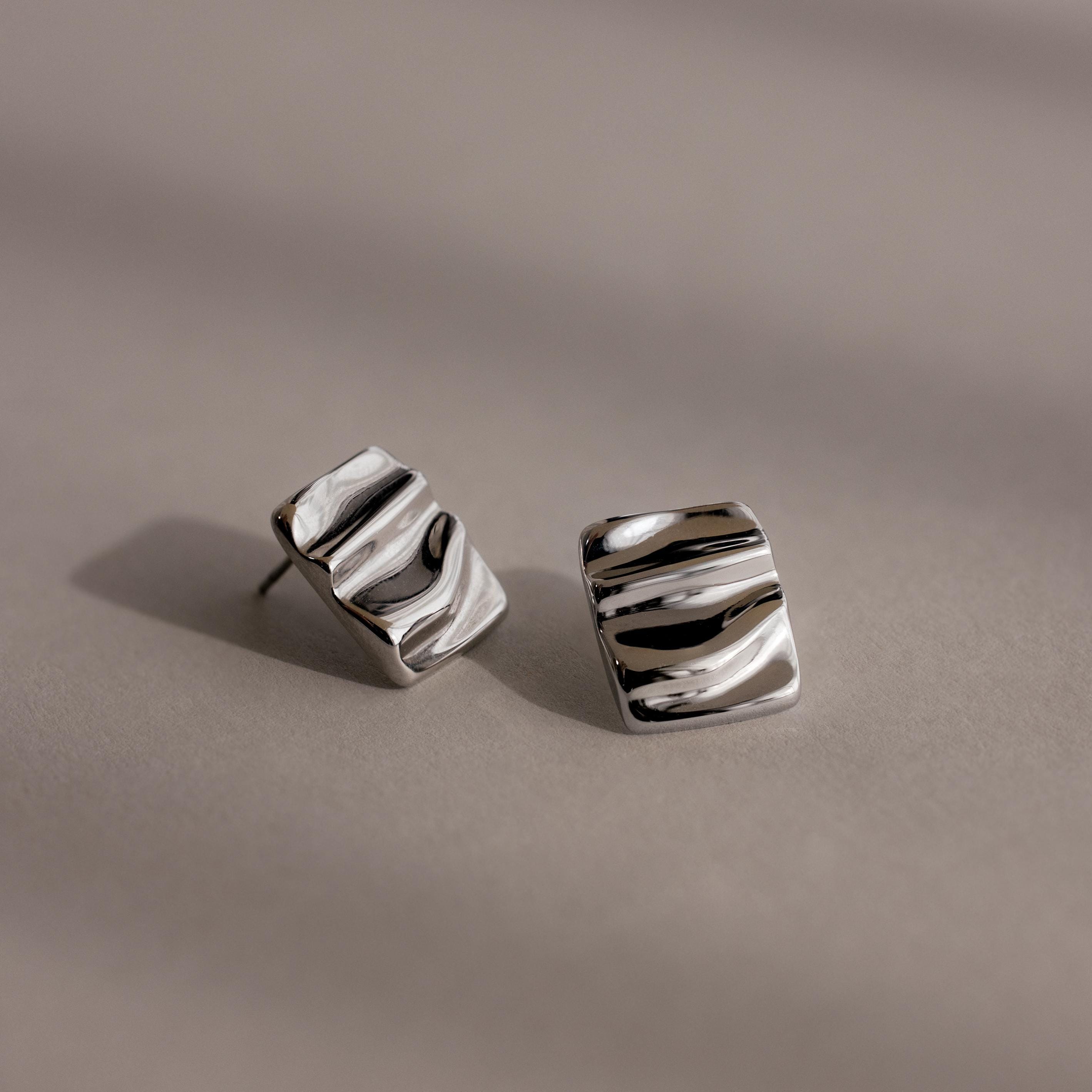 Bold Square Wave Studs in silver feature a wavy, textured surface for a unique touch. Displayed on a neutral background, these minimalist earrings add modern elegance to any luxury jewelry collection.