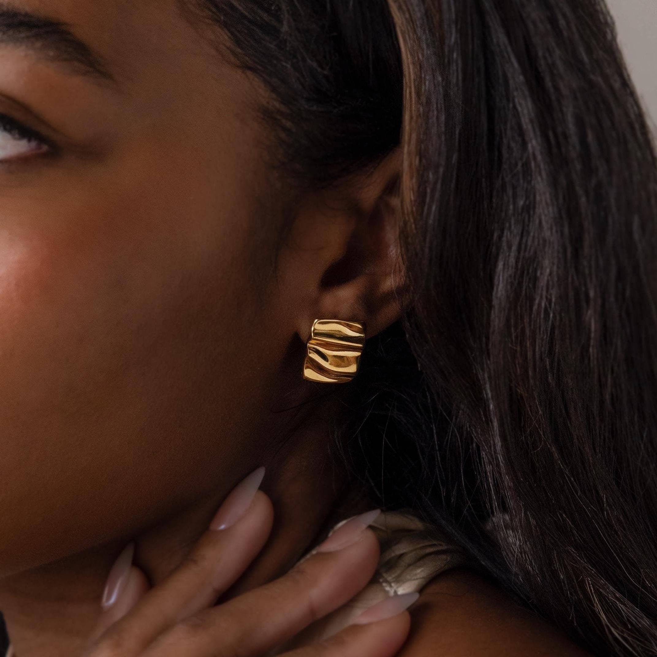 Close-up of a woman's ear wearing Bold Square Wave Studs—chunky gold geometric minimalist earrings. Her long dark hair enhances the striking touch of luxury jewelry.