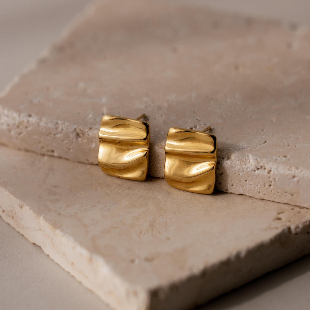 Bold Square Wave Studs, featuring a wavy texture and minimalist design, rest on beige stone slabs—luxury earrings from our jewelry collection.