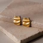 Bold Square Wave Studs, featuring a wavy texture and minimalist design, rest on beige stone slabs—luxury earrings from our jewelry collection.