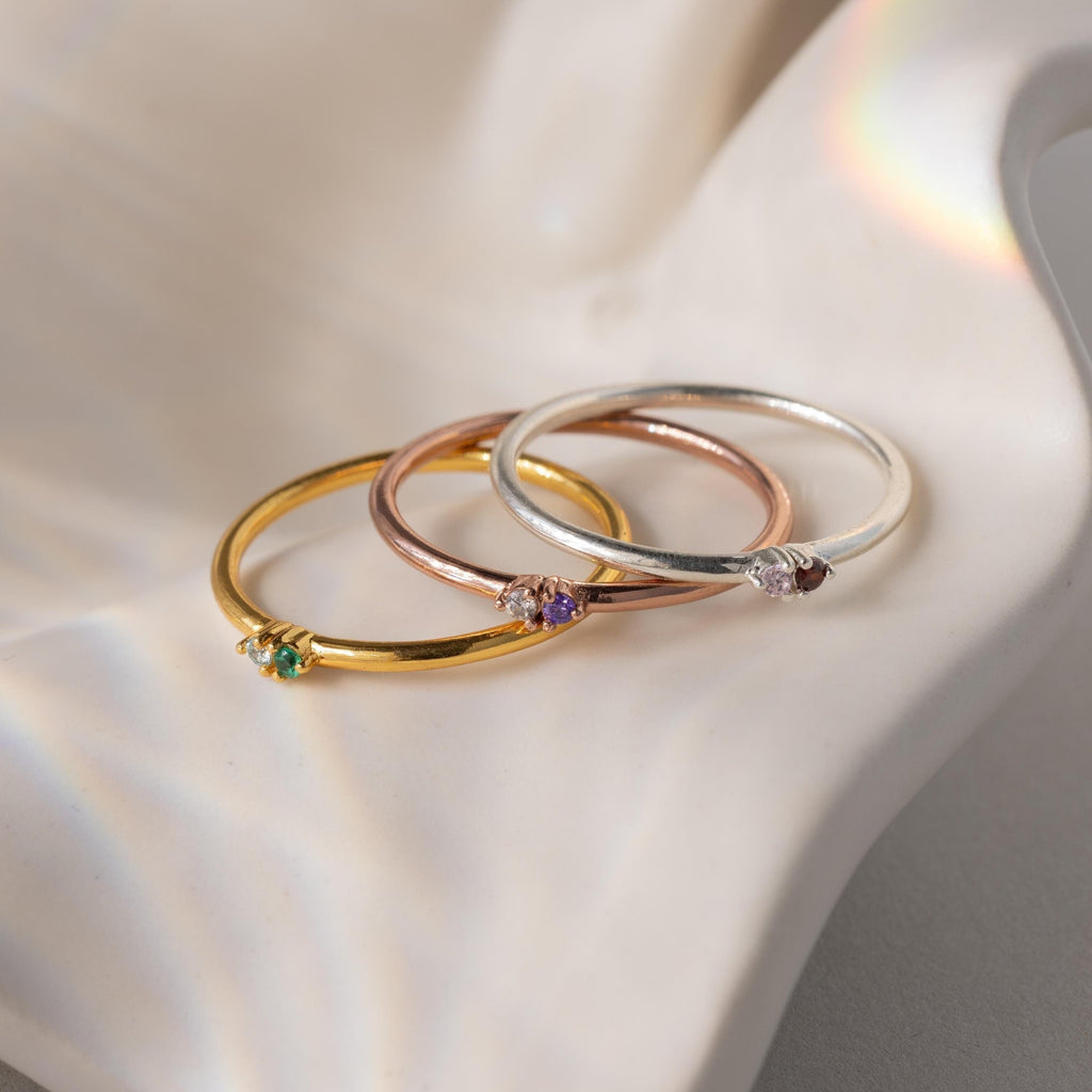 The Mini Toi et Moi Birthstone Rings in gold, rose gold, and silver each feature a small gemstone and are displayed elegantly on a wavy white surface.