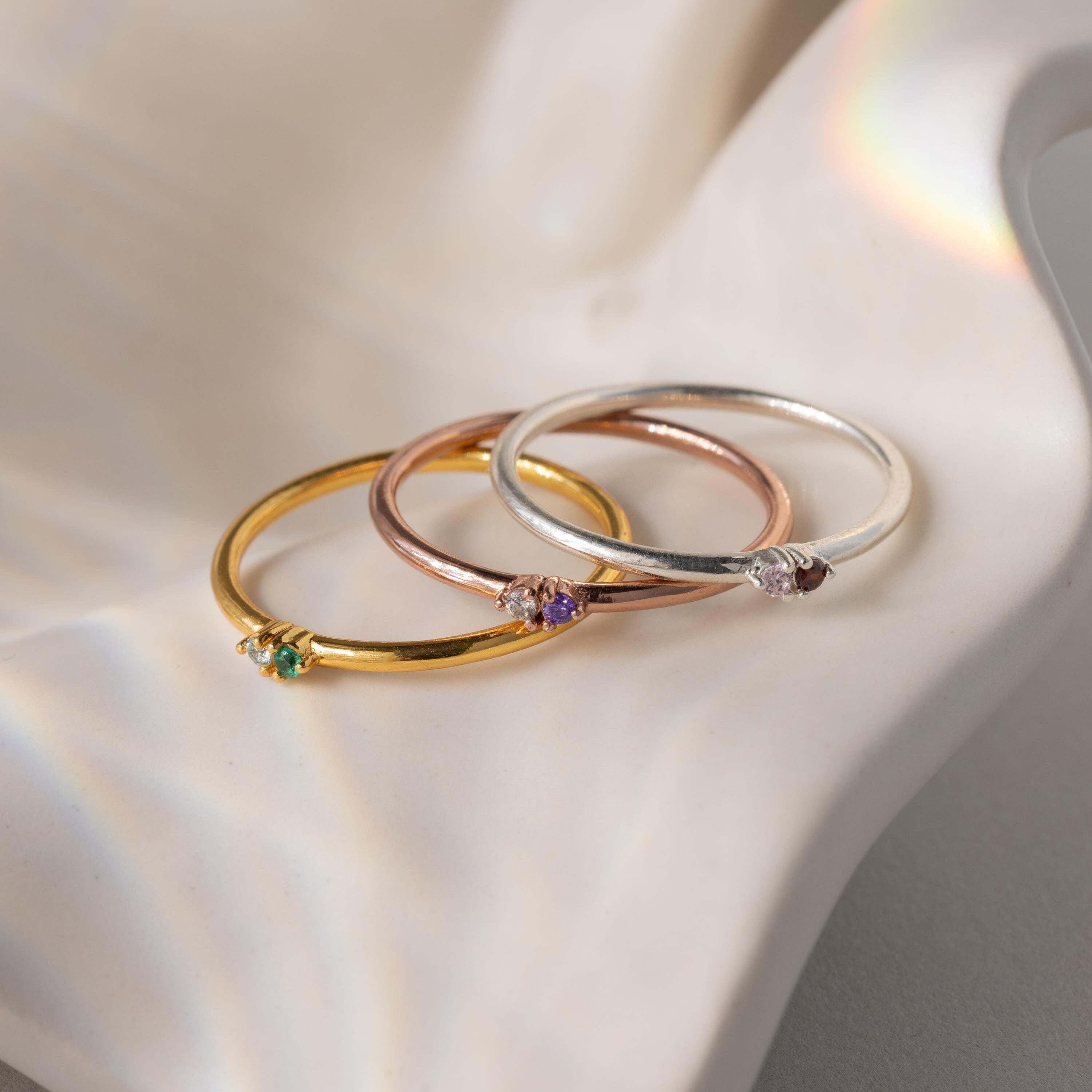 The Mini Toi et Moi Birthstone Rings in gold, rose gold, and silver each feature a small gemstone and are displayed elegantly on a wavy white surface.