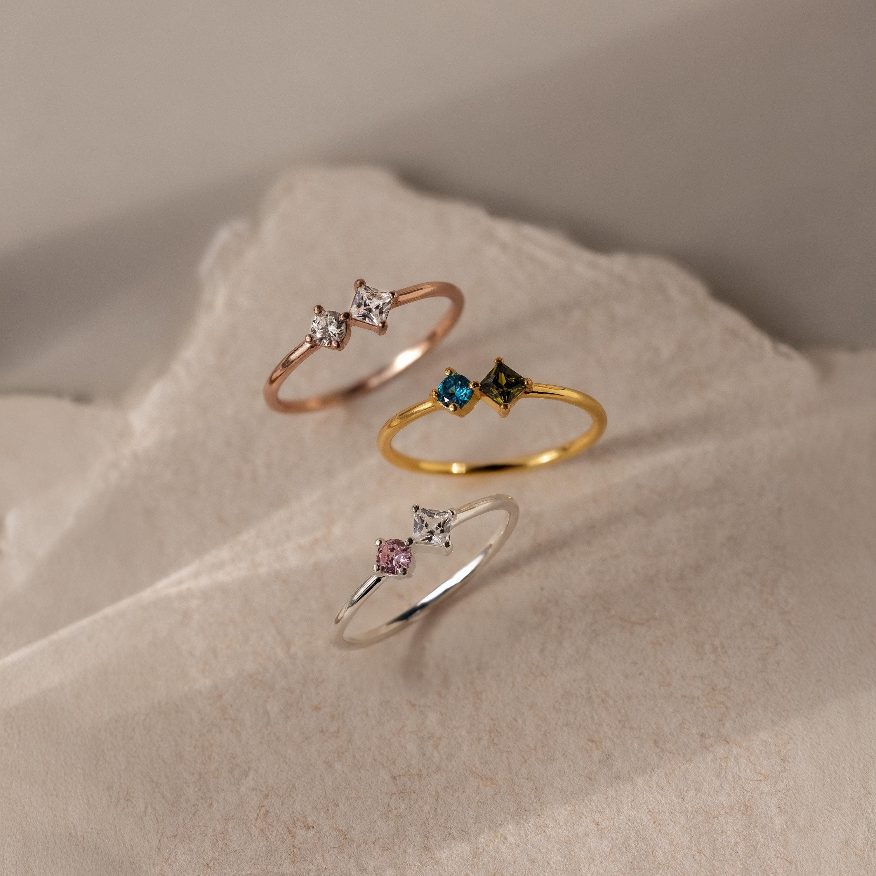 The Toi et Moi Princess Birthstone Ring features a customizable design with colorful gemstones, displayed on textured, neutral-toned surfaces.