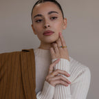 Woman in a cream ribbed top and brown blazer, showcasing gold rings—including the Toi et Moi Emerald Birthstone Ring—on her fingers against a neutral background.