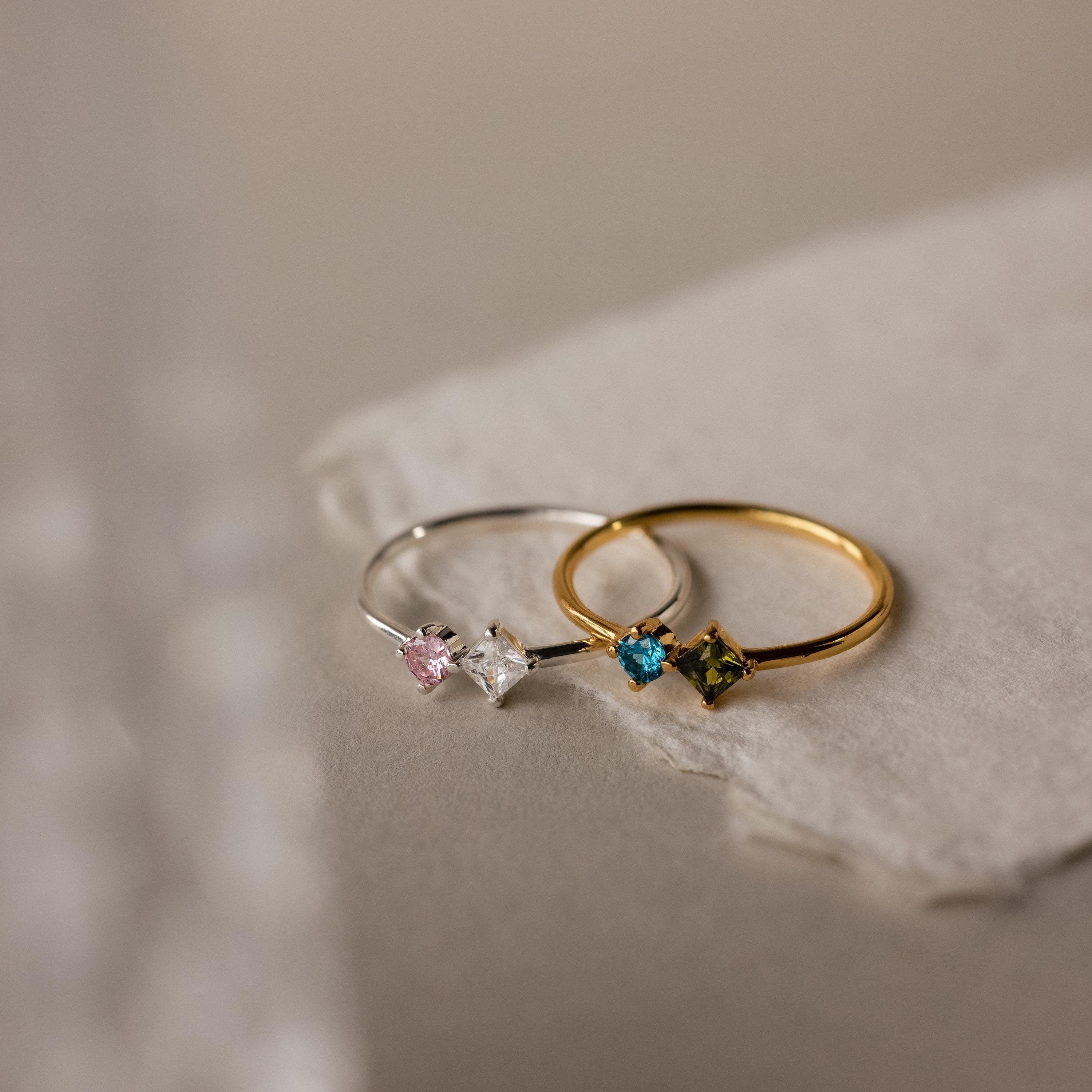 The Toi et Moi Princess Birthstone Ring features customizable colored gemstones—pink, clear, blue, and green—set in a delicate design. Perfect for stacking or as a unique birthstone ring on a textured white surface.