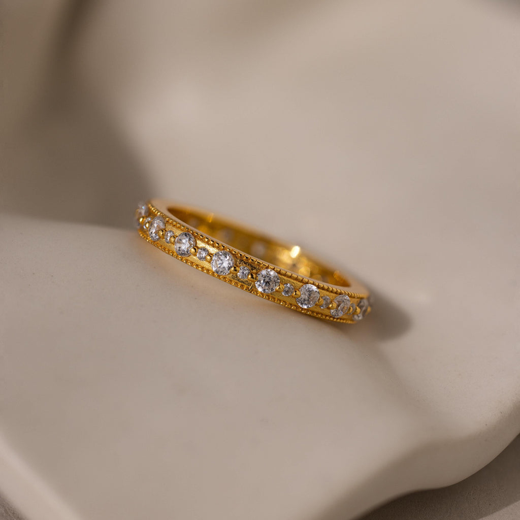 The Diamond Eternity Ring, a gold stackable band featuring a row of small round diamonds, is displayed on a soft beige surface.