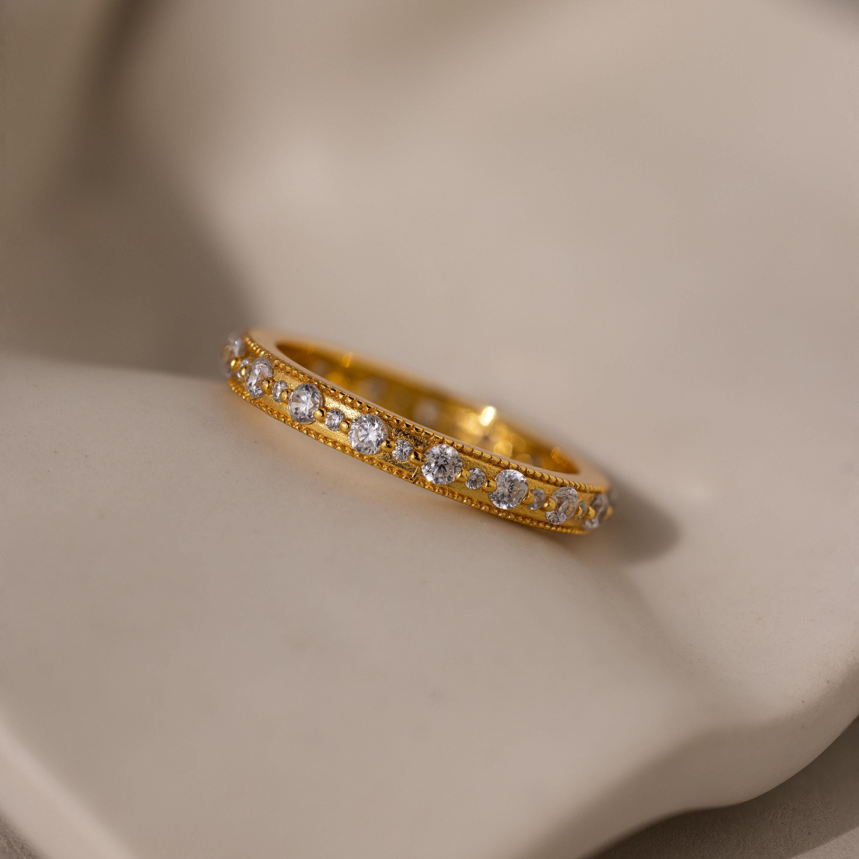 The Diamond Eternity Ring, a gold stackable band featuring a row of small round diamonds, is displayed on a soft beige surface.