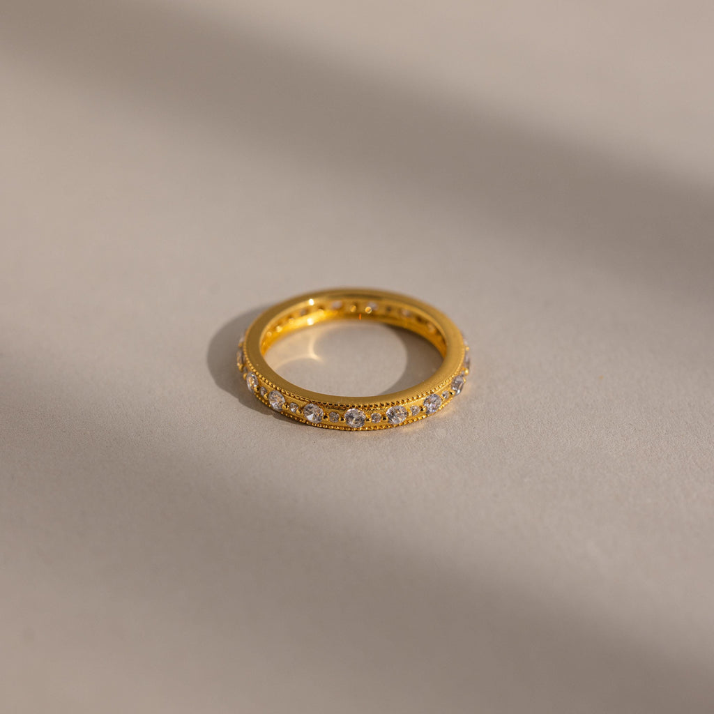 A Diamond Eternity Ring featuring small diamonds around the gold band, displayed on a softly lit neutral surface.