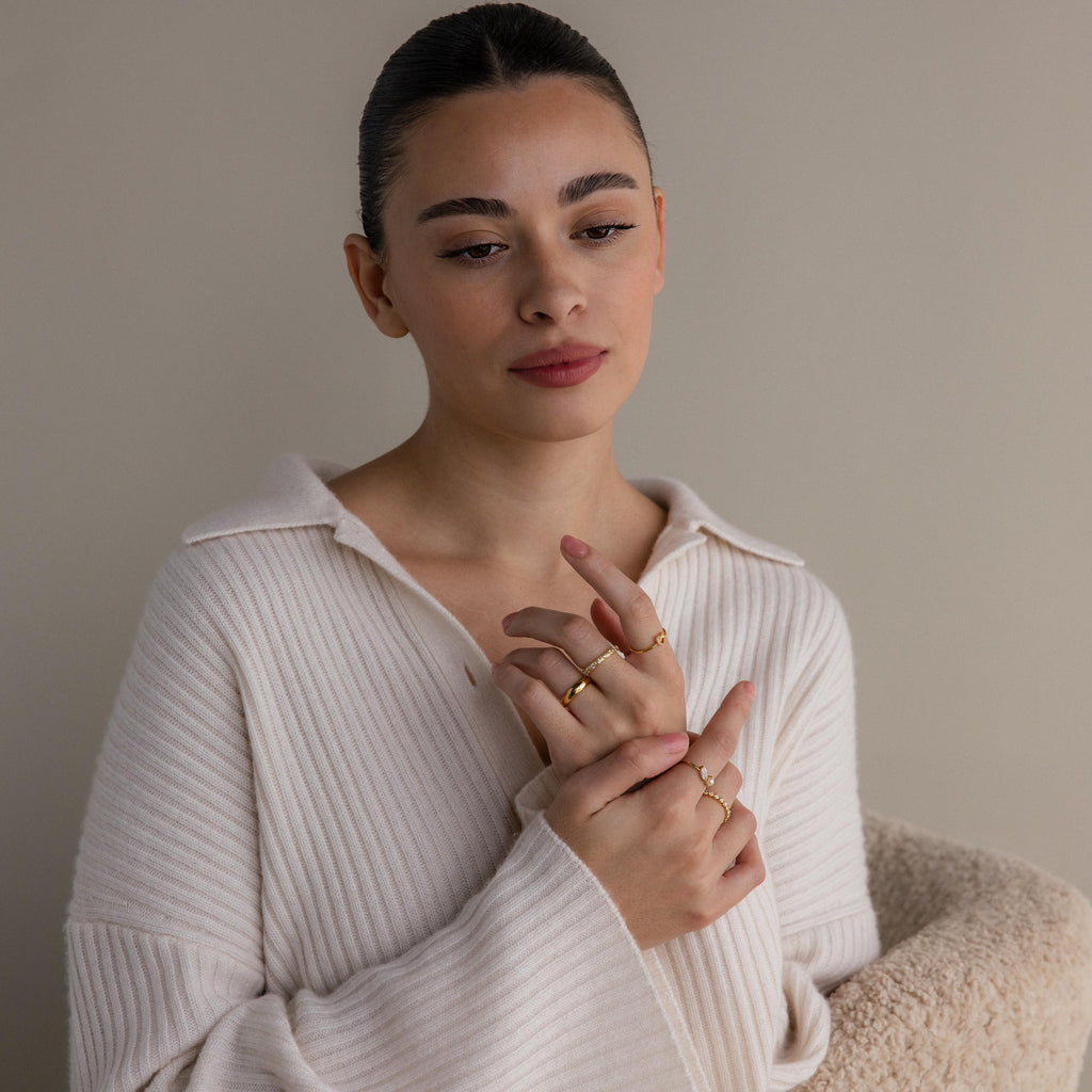 A woman wearing a cream ribbed sweater displays the Diamond Eternity Ring along with other gold stackable rings on her fingers, set against a neutral background.