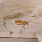 The Diamond Eternity Ring, a stackable gold band adorned with small diamonds, is displayed on a textured cream surface with a soft fabric background.